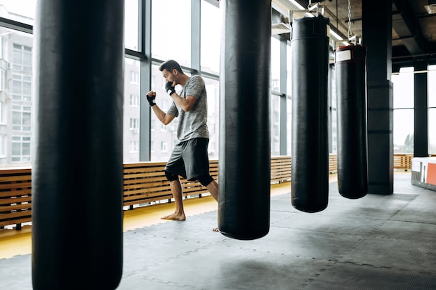 Der dunkelhaarige Typ im grauen T-Shirt und schwarzen Shorts steht hinter dem hängenden Boxsack neben Panoramafenstern in der Boxhalle.