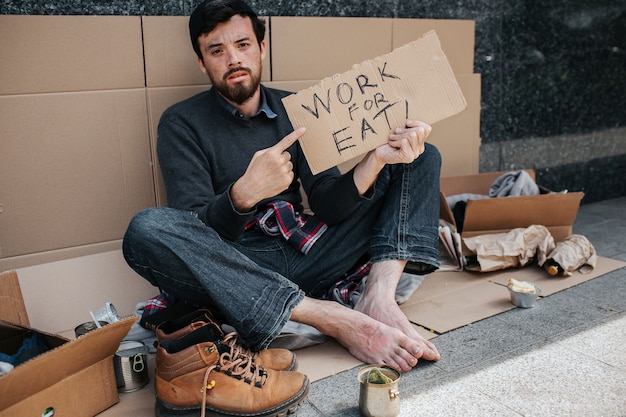Der dunkelhaarige und bärtige Obdachlose sitzt und zeigt auf ein Stück Pappe, auf dem „Arbeit für Essen“ steht