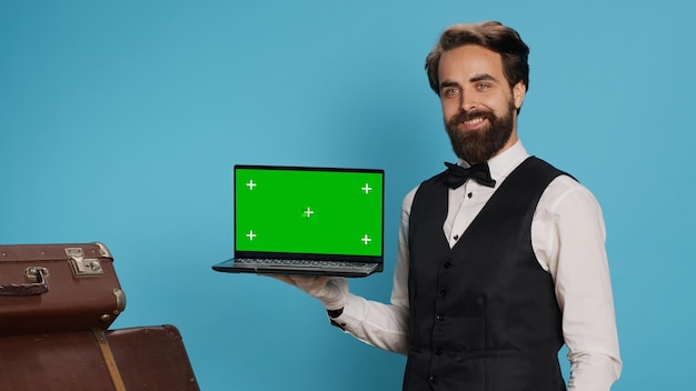 Der elegante Hotelpage zeigt Greenscreen auf dem Laptop im Studio, während er weiße Handschuhe neben dem Koffer der Gäste trägt. Luxuriöser, stilvoller Portier, der in einem modernen Hotel arbeitet und isolierte Modelldisplays präsentiert.