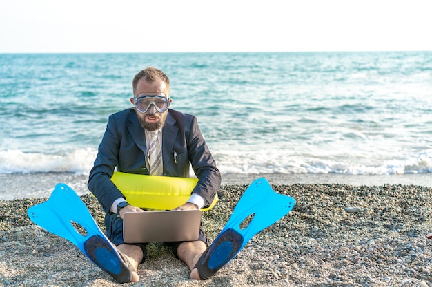 Der erfolgreiche Geschäftsmann, der Werkzeuge schnorchelnd trägt, arbeitet an dem Strand mit Laptop