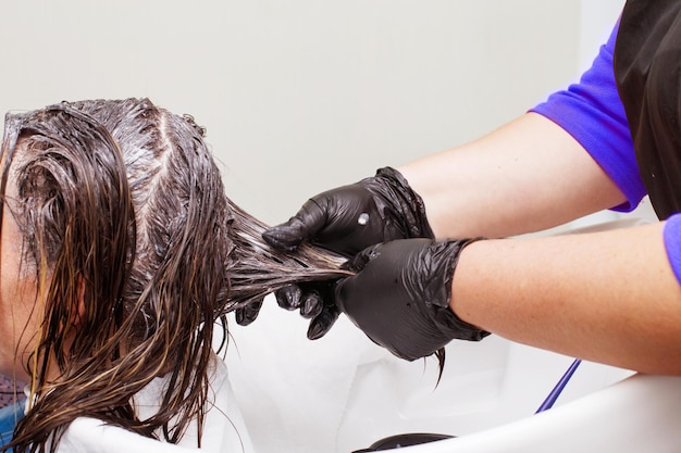 Der Friseur in schwarzen Handschuhen malt im Schönheitssalon die Haare einer brünetten Frau