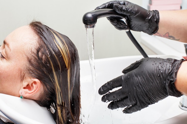 Der Friseur in schwarzen Handschuhen wäscht die Haare der brünetten Frau im Schönheitssalon