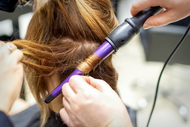 Der Friseur macht mit dem Lockenstab im Friseursalon aus nächster Nähe Locken aus langen braunen Haaren