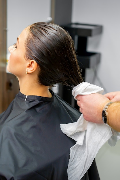 Der Friseur trocknet das gewaschene Haar mit einem Handtuch an eine schöne junge Brünette in einem Schönheitssalon