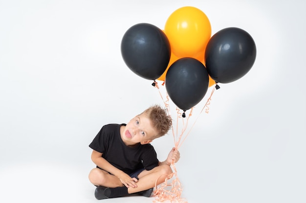 Der fröhliche, fröhliche Halloween-Kinderjunge im schwarzen T-Shirt zeigt seine Zunge, die schwarze und orangefarbene Luftballons hält
