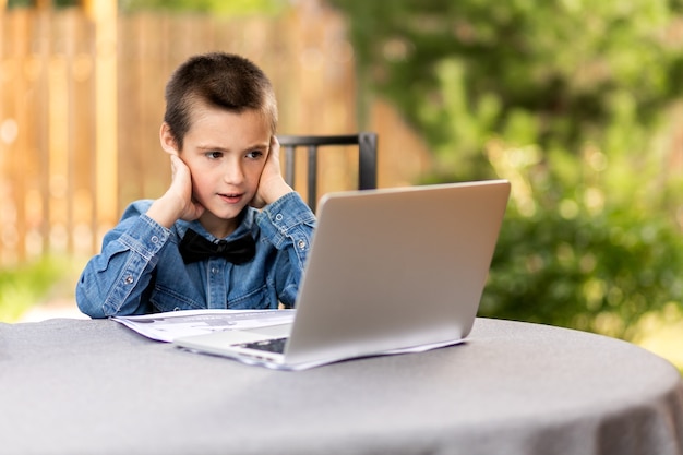 Der fröhliche Schuljunge nimmt zu Hause im Garten über einen Laptop Unterricht. Online-Kurse für Kinder