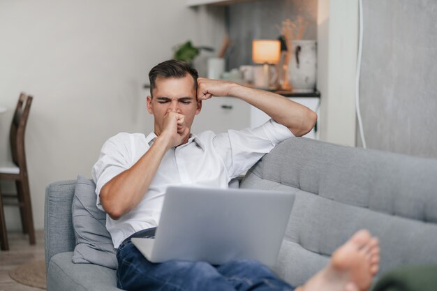 Der gähnende junge Mann mit einem Laptop ruht sich im Wohnzimmer aus.