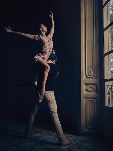 Der ganze Körper einer anmutigen Ballerina mit erhobenem Arm sitzt auf der Schulter eines barfüßigen männlichen Tänzers, während er in einem dunklen Raum tanzt