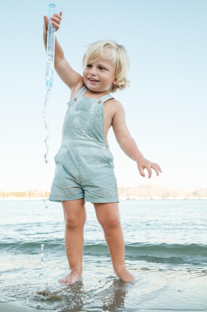 Der ganze Körper eines fröhlichen kleinen blonden Kindes in lässiger Kleidung, das während der Sommerferien eine Seifenblasenflasche im Meer am Strand leert