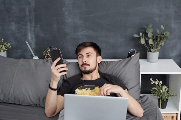 Der gelangweilte junge Student liegt im Bett und isst Chips, während er auf dem Smartphone im Internet surft. Während der Quarantäne verschwendet er zu Hause Zeit