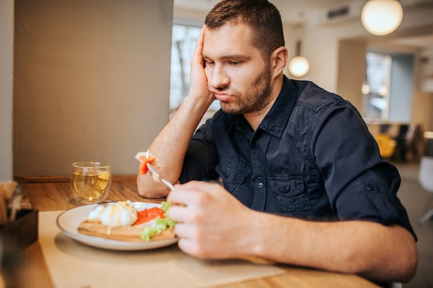 Der gelangweilte und traurige Mann sitzt am Tisch und im Café. Er hält ein Stück Gemüse an der Gabel. Der Mensch schaut es an und atmet aus.