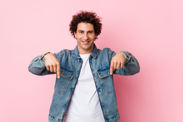 Der gelockte reife Mann, der eine Denimjacke gegen rosa Wand trägt, zeigt unten mit den Fingern, positives Gefühl.