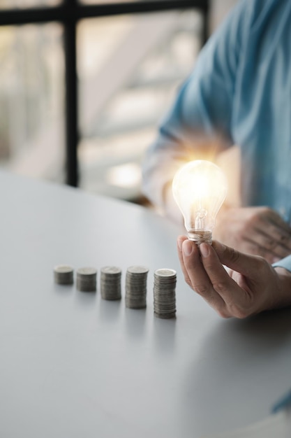 Der Geschäftsmann hält eine leuchtende Glühbirne auf dem höchsten Münzstapel. Das Platzieren von Münzen in einer Reihe von niedrig nach hoch ist vergleichbar mit dem Sparen von Geld, um mehr zu wachsen Geld sparende Ideen für Investitionen in Fonds