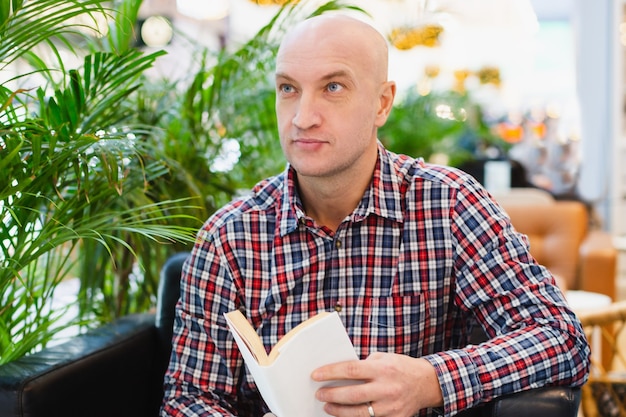 Der glatzköpfige Europäer in einem rot karierten Hemd und Blue Jeans sitzt in einem Sessel in einer hellen Wohnung mit vielen grünen Pflanzen und liest ein Buch in einem entspannenden Innenraum