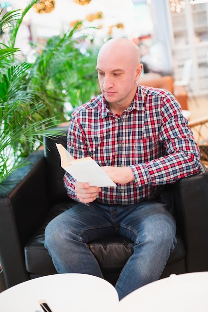 Der glatzköpfige Europäer in einem rot karierten Hemd und Blue Jeans sitzt in einem Sessel in einer hellen Wohnung mit vielen grünen Pflanzen und liest ein Buch in einem entspannenden Innenraum