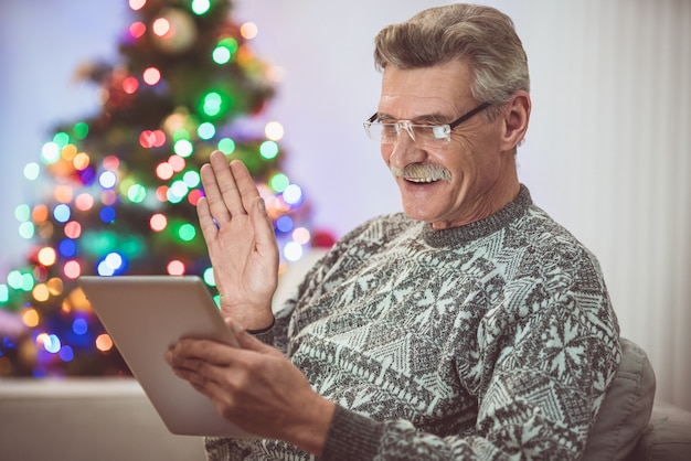 Der glückliche alte Mann mit einem Tablet macht einen Videoanruf in der Nähe des Weihnachtsbaums