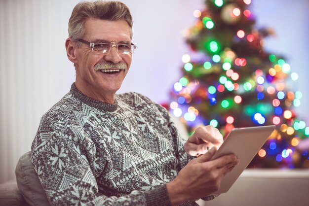 Der glückliche alte Mann mit einem Tablet sitzt neben dem Weihnachtsbaum