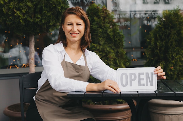 Der glückliche Besitzer eines Cafés in einer Schürze hält ein offenes Schild in der Nähe des Cafés und schaut in die Kamera. weibliche Kellnerin.