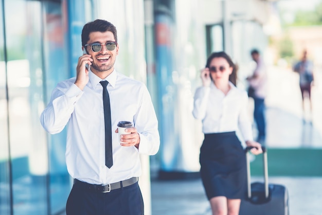 Der glückliche Geschäftsmann mit Sonnenbrille telefoniert in der Nähe der Frau mit einem Koffer