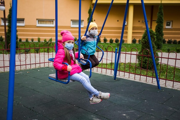 Der glückliche kleine Junge mit medizinischer Maske sitzt auf einer Holzkonstruktion auf dem Spielplatz Das kleine Mädchen spielt hinter ihm