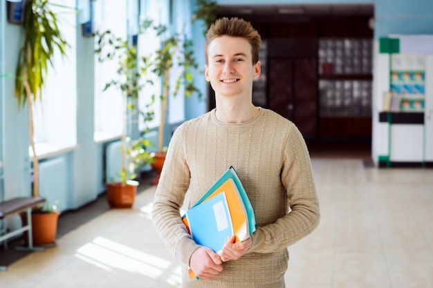 Der glückliche männliche Student, der im Campus am sonnigen Tag hält Bücher, Übungsbücher steht, glücklich, in der Universität zu studieren und hohe akademische Leistung zu haben