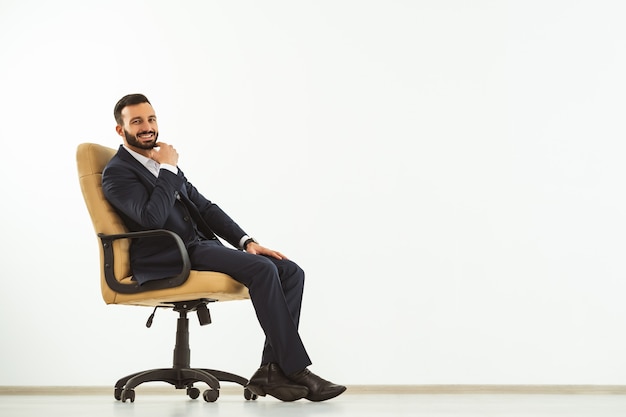 Der glückliche Mann, der auf einem Bürostuhl auf dem weißen Wandhintergrund sitzt