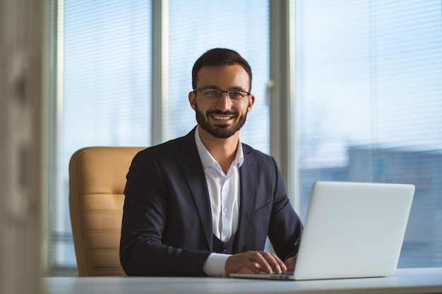 Der glückliche Mann mit Brille, der mit einem Laptop am Schreibtisch arbeitet