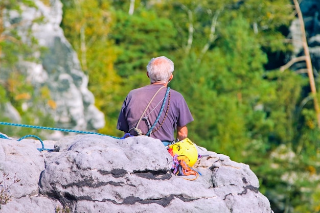 Der grauhaarige Mann sitzt auf einem Felsen vor einem Hintergrund aus Wald und Bergen