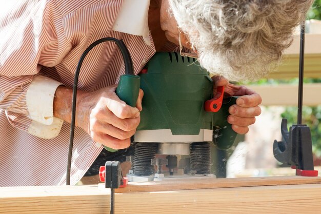 Foto der grauhaarige zimmermann hält in seinen händen ein werkzeug, eine grüne fräsmaschine, und schneidet es auf einem frischen holzbrett aus, während er an einem sommerhaus baut. tischlerei und holzverarbeitung
