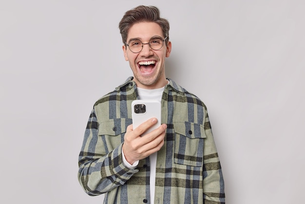 Der gutaussehende Brunet-Mann trägt eine runde Brille und ein kariertes Hemd verwendet ein Smartphone, sendet SMS, scrollt ins Internet und posiert fröhlich vor einer grauen Studiowand. Konzept für Menschen und moderne Technologien