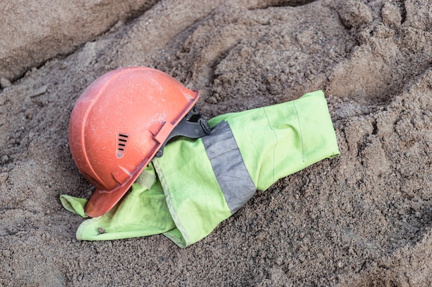 Der Helm und die Signalweste eines orangefarbenen Bauarbeiters ruhen auf einem Sandhaufen, um Beton vorzubereiten. Mittagspause auf einer Baustelle.