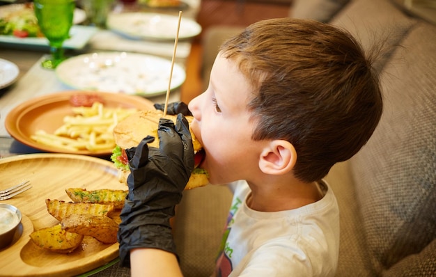 Der hübsche kleine Junge isst Burger in schwarzen Gummihandschuhen