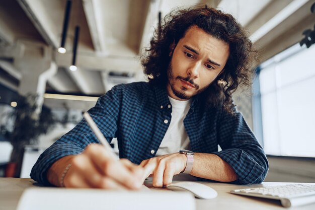 Der hübsche lockige Kerl macht sich am Arbeitstisch im Großraumbüro Notizen