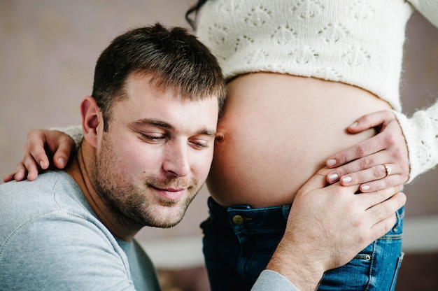 Der hübsche Mann hört dem Bauch seiner schönen schwangeren Frau zu und lächelt.