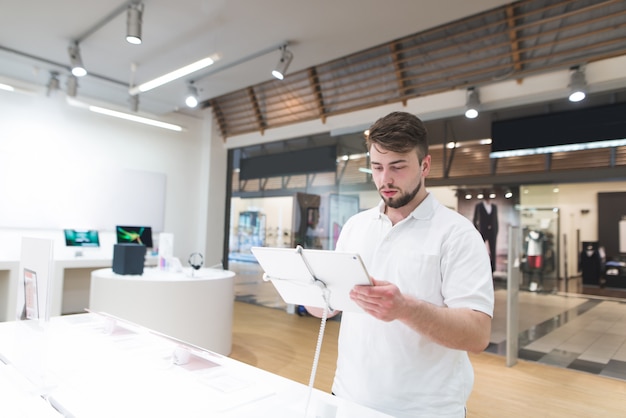 Der hübsche Mann steht mit einer Tablette in den Händen in einem modernen Tech-Laden