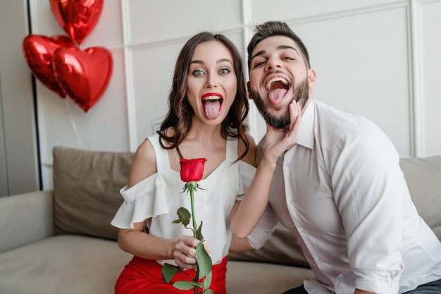Der hübsche Paarmann und -frau, die Zunge mit Rotrose und Herz zeigen, formten Ballone zu Hause auf Couch