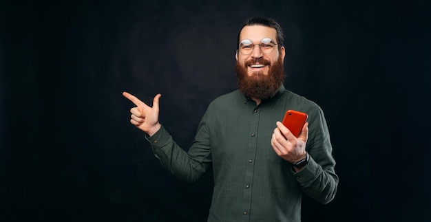 Der intelligente bärtige Mann hält ein rotes Telefon, während er auf den Hintergrund zeigt Kopierbereich über schwarzem Hintergrund in einem Studio
