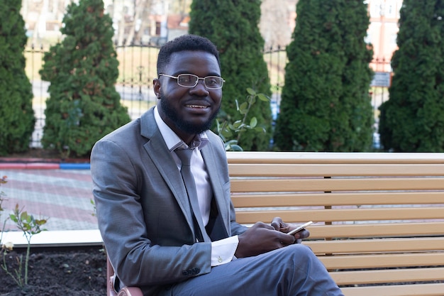 Der junge afrikanische Geschäftsmann, der auf Stadtbank mit Telefon, Tablette lächelt