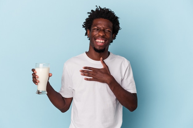 Der junge Afroamerikaner, der ein Glas Milch auf blauem Hintergrund hält, lacht laut und hält die Hand auf der Brust.