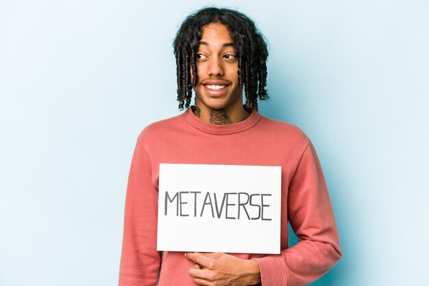 Der junge Afroamerikaner, der ein Metaverse-Plakat isoliert auf blauem Hintergrund hält, sieht beiseite und lächelt fröhlich und angenehm