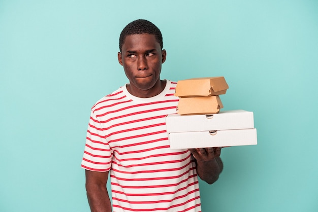 Der junge Afroamerikaner, der Pizza und Burger einzeln auf blauem Hintergrund hält, verwirrt, fühlt sich zweifelhaft und unsicher.