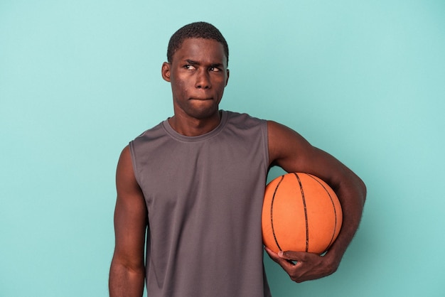 Der junge afroamerikanische Mann, der isoliert auf blauem Hintergrund Basketball spielt, fühlt sich verwirrt und unsicher