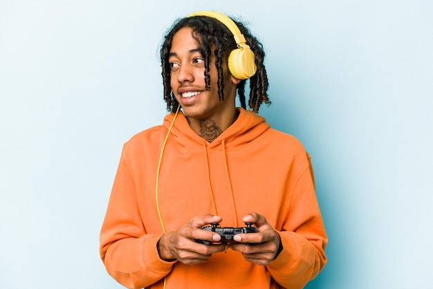 Der junge afroamerikanische Mann, der mit einem Videospiel-Controller spielt, der isoliert auf blauem Hintergrund steht, sieht beiseite und lächelt fröhlich und angenehm