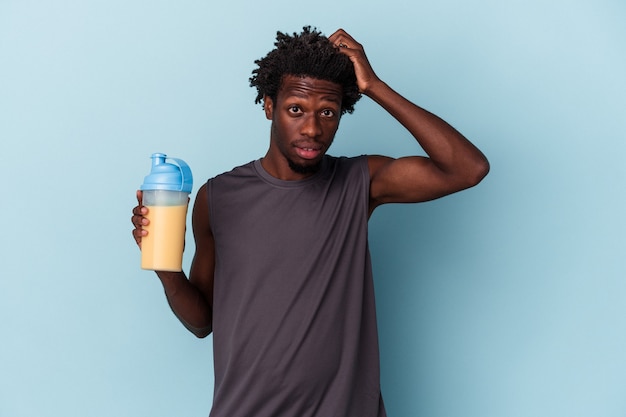 Der junge afroamerikanische Mann, der Protein-Milchshake isoliert auf blauem Hintergrund hält, ist schockiert, sie hat sich an ein wichtiges Treffen erinnert.