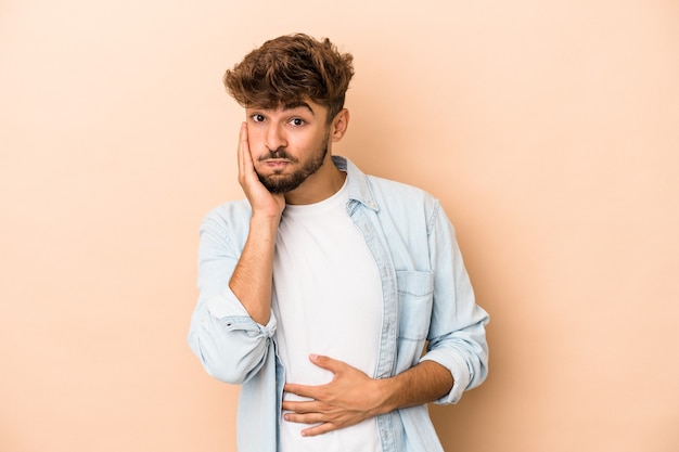 Der junge arabische Mann, der auf beigem Hintergrund isoliert ist, bläst Wangen, hat müden Ausdruck. Gesichtsausdruck Konzept.