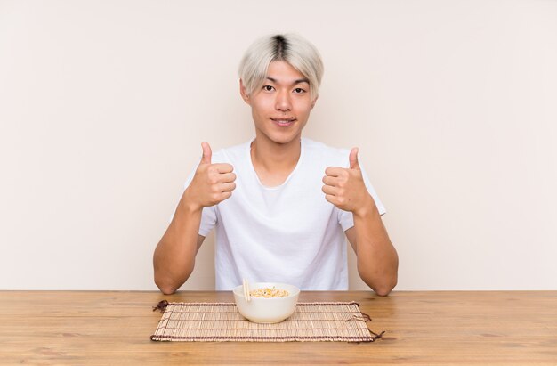 Der junge asiatische Mann mit dem Ramen in einer Tabelle gebend Daumen up Geste