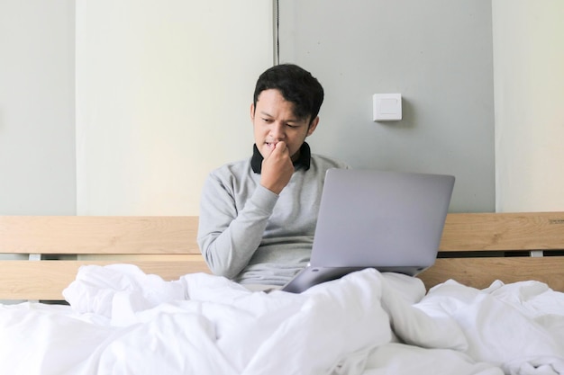 Der junge asiatische Mann mit einem Laptop im Bett ist verwirrt und stresst das Konzept der Arbeit von zu Hause aus