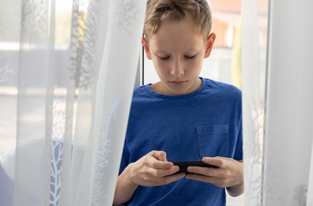 Der Junge benutzt das Smartphone, während er zu Hause auf der Fensterbank sitzt