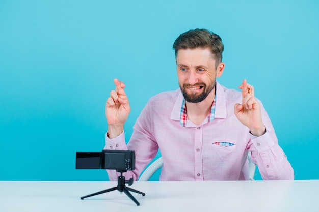 Der junge Blogger-Mann posiert vor seiner Mini-Kamera, indem er die Finger auf blauem Hintergrund kreuzt