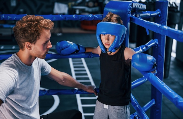 Der junge Boxtrainer hilft dem kleinen Jungen in Schutzkleidung auf dem Ring zwischen den Runden.
